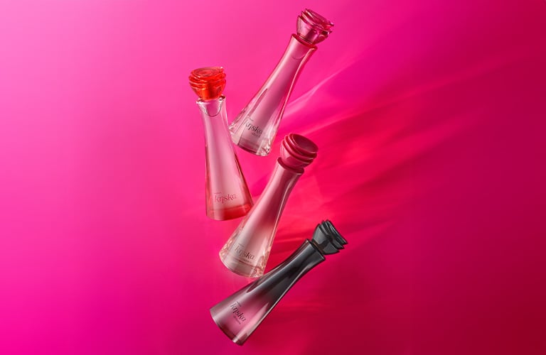 Foto de cuatro frascos de perfume Kriska de Natura, en tonos rosa, rojo y negro, con tapas en forma de flor, dispuestos sobre un fondo rosa vibrante.